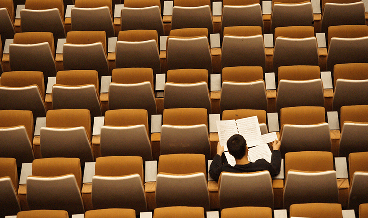 Un hombre sale leyendo unos apuntes, en un asiento de un auditorio vacío de butacas de color marrón clarito